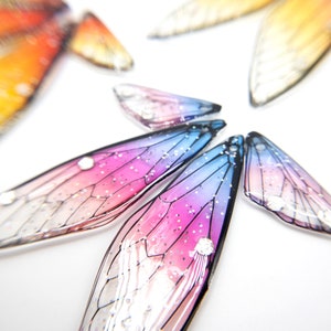 May include: A set of iridescent butterfly wings with a pink, blue, and white gradient. The wings are made of a clear material with a glitter finish.