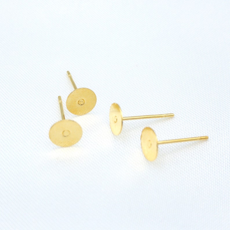 May include: Four gold-colored earring studs with flat circular pads for attaching components. Each stud features a thin, straight post extending from the center of the back of the circular pad, designed for insertion into an earlobe piercing.