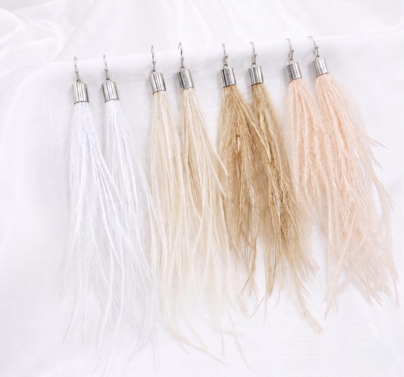 Puede incluir: Una colecci&oacute;n de pendientes largos colgantes con borlas en forma de plumas en varios colores neutros. Cada pendiente presenta una tapa y un gancho de metal plateado. Los colores var&iacute;an del blanco al beige y marr&oacute;n claro, creando una est&eacute;tica suave y elegante.