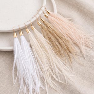 Puede incluir: Pendientes de plumas en blanco, crema, beige y rosa rubor. Cada pendiente tiene una tapa y un gancho de metal dorado. Los pendientes se exhiben sobre un plato blanco y una tela de color neutro.