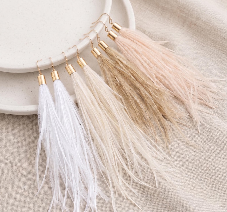 Puede incluir: Pendientes de plumas en blanco, crema, beige y rosa rubor. Cada pendiente tiene una tapa y un gancho de metal dorado. Los pendientes se exhiben sobre un plato blanco y una tela de color neutro.