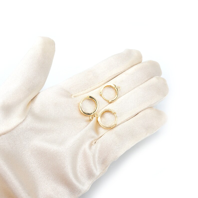 Op de afbeelding: Drie kleine, cirkelvormige goudkleurige oorringen, elk voorzien van een scharnierend sluitingsmechanisme en een minuscuul ge&iuml;ntegreerd lusje aan de basis voor het bevestigen van extra sieraadonderdelen. De ringen hebben een gepolijste afwerking.