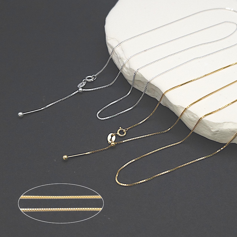 Puede incluir: Dos collares de cadena ajustables, uno plateado y otro dorado, se encuentran sobre una superficie blanca. Las cadenas son de estilo caja y tienen una peque&ntilde;a bola al final del deslizador ajustable.