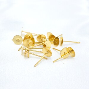 May include: A collection of gold-colored metal earring posts, each featuring a flat circular pad at the top for attaching decorative elements and a thin, straight pin extending from the center, designed for use as jewelry making components.