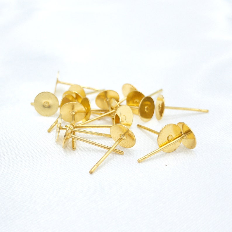 May include: A collection of gold-colored metal earring posts, each featuring a flat circular pad at the top for attaching decorative elements and a thin, straight pin extending from the center, designed for use as jewelry making components.