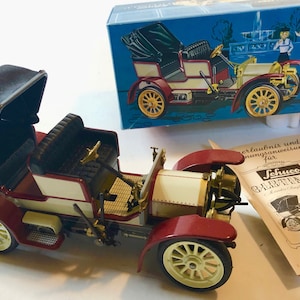 May include: A vintage toy car model with a black convertible top, cream-colored body, and maroon fenders. The model has gold-colored wheels and accents. A blue box with a vintage car illustration and a pamphlet are also present.