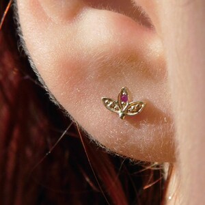May include: A close-up of a person's ear with a small gold lotus flower earring with a red gemstone in the center.