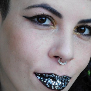 May include: Close-up of a person's face with dramatic makeup. The eyes are accentuated with black eyeliner and gold eyeshadow. The lips are painted black and adorned with silver star-shaped glitter. A silver nose ring is also visible.