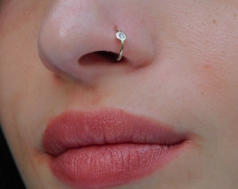 May include: Close-up of a person's face with a small gold nose ring with a clear gemstone. The person is wearing pink lipstick.