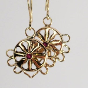 May include: A pair of gold-colored earrings. Each earring features a circular design with a central red gemstone and a floral-like outer edge. The earrings are suspended from a simple hook.