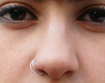 May include: A close-up of a woman's face with dark hair, dark eye makeup, and a nose ring. She is wearing earrings with a circular design.