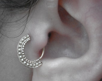 Könnte beinhalten: Ein silberner Septumring mit einem halbmondförmigen Design. Der Ring hat eine Reihe kleiner, gleichmäßig verteilter Perlen entlang seiner gebogenen Oberfläche. Der Schmuck wird in einem durchbohrten Ohr getragen.