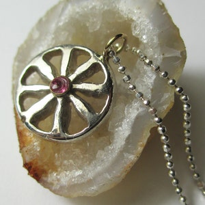 May include: A silver pendant necklace featuring a circular design with a central pink gemstone. The pendant has an openwork pattern and hangs from a delicate silver chain. The necklace is displayed on a white and brown surface.