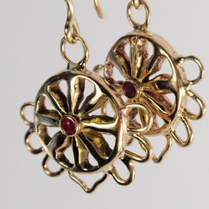 May include: A pair of gold-colored earrings with a circular design. Each earring features a central red gemstone and an openwork pattern with radiating lines. The earrings have a hook closure and a floral-like outer edge.