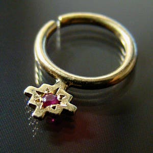 May include: A gold-colored ring with a star-shaped charm. The charm features a small, round, red gemstone at its center. The ring is set against a dark, reflective surface, highlighting its polished finish.