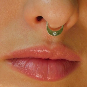 Puede incluir: Primer plano de la nariz de una persona con un anillo de septum de oro con una pequeña piedra preciosa transparente.