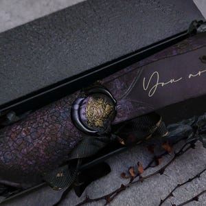 Puede incluir: Caja de invitación rectangular de color púrpura oscuro con un patrón agrietado. La caja está sellada con un sello de cera con una rosa dorada. Las palabras "You are INVITED!" están escritas en oro.