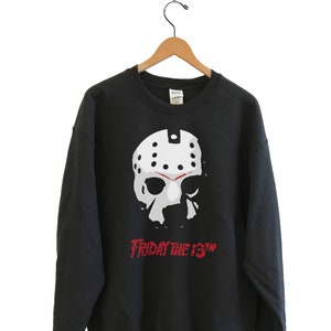 Op de afbeelding: Zwart sweatshirt met een wit hockeymasker ontwerp en de rode tekst "FRIDAY THE 13TH". Het kledingstuk hangt aan een houten kledinghanger.