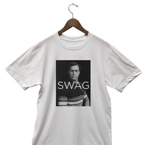 May include: White t-shirt with a black and white image of a man wearing glasses and a striped shirt. The word "SWAG" is printed in white letters below the image. The t-shirt is hanging on a wooden hanger.