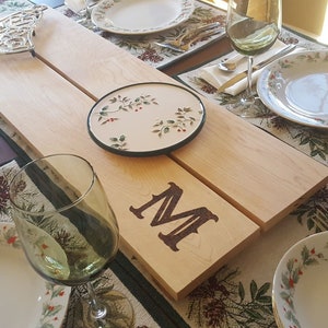 May include: A wooden serving board with the letter "M" burned into the wood, set on a table with place settings. The table is set with plates, silverware, and wine glasses. A decorative trivet and a small plate are also on the board.