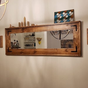 May include: A rectangular wooden mirror with a brown stained finish and black metal corner brackets. The mirror is mounted on a wall above a shelf with decorative items.