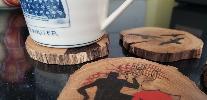 May include: A white ceramic mug with a blue typewriter design and the word "EWRITER" rests on a wooden coaster. Additional wooden coasters with painted designs of birds and a figure are also visible.