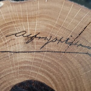 May include: A cross-section of a tree trunk with visible growth rings and a handwritten signature in black ink. The wood has a light brown color with darker brown lines and a rough, natural edge.