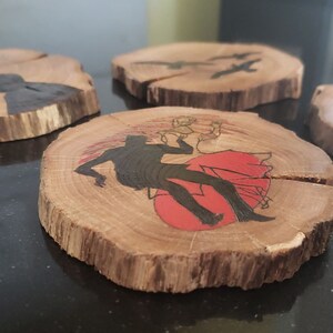 May include: Set of four wooden coasters with unique designs. Each coaster is a natural wood slice. One features a silhouette of a person with a red sun, another has birds, and the others have abstract black shapes.