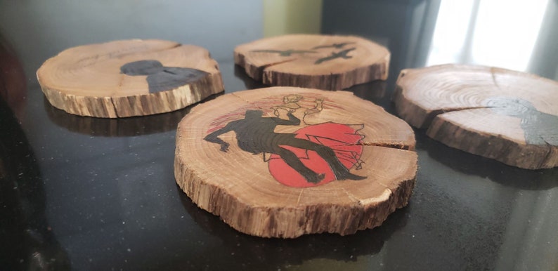 May include: Set of four wooden coasters with unique designs. Each coaster is a natural wood slice. One features a silhouette of a person with a red sun, another has birds, and the others have abstract black shapes.