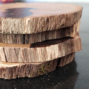 May include: A stack of four round wooden coasters. Each coaster is made from a cross-section of a tree branch, displaying the natural wood grain and bark. The coasters have a rustic, natural appearance, with variations in color and texture.