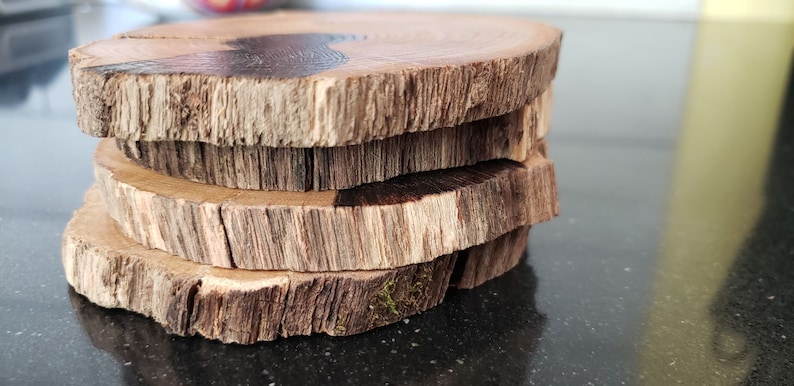 May include: A stack of four round wooden coasters. Each coaster is made from a cross-section of a tree branch, displaying the natural wood grain and bark. The coasters have a rustic, natural appearance, with variations in color and texture.