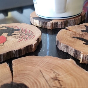 May include: Set of four wooden coasters with unique designs. One coaster features a black silhouette of a figure with a red background, another has three black birds, and the others have abstract designs. The coasters are made from natural wood slices.