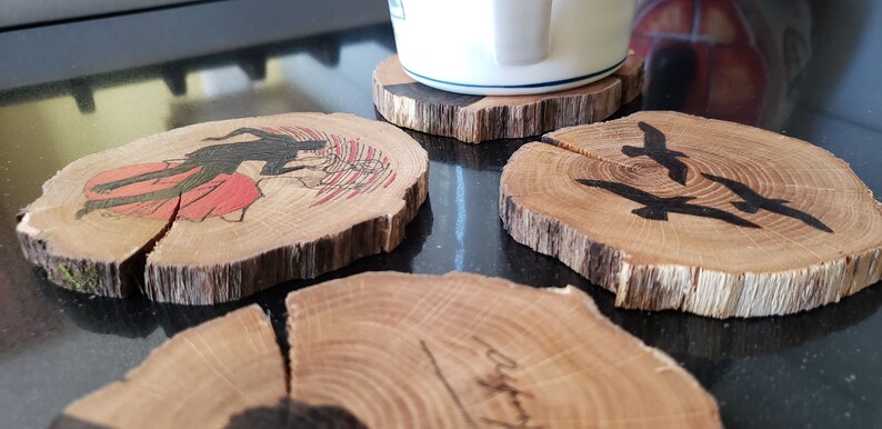 May include: Set of four wooden coasters with unique designs. One coaster features a black silhouette of a figure with a red background, another has three black birds, and the others have abstract designs. The coasters are made from natural wood slices.