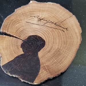 May include: Wooden coasters with a natural wood grain. One coaster features a dark silhouette of a person and a signature. The other coaster has a red and black illustration. These coasters are perfect for home decor.