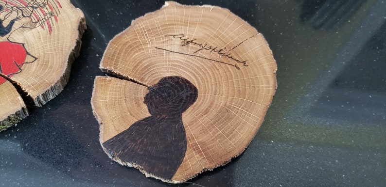 May include: Wooden coasters with a natural wood grain. One coaster features a dark silhouette of a person and a signature. The other coaster has a red and black illustration. These coasters are perfect for home decor.