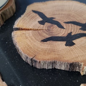 May include: Wooden coasters with a natural edge. One coaster features three black painted birds in flight. The coasters are made from a cross-section of a tree branch, with visible wood grain and a light brown color.