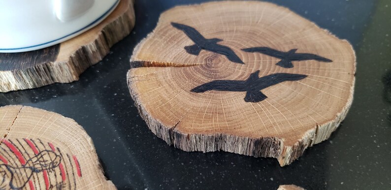 May include: Wooden coasters with a natural edge. One coaster features three black painted birds in flight. The coasters are made from a cross-section of a tree branch, with visible wood grain and a light brown color.