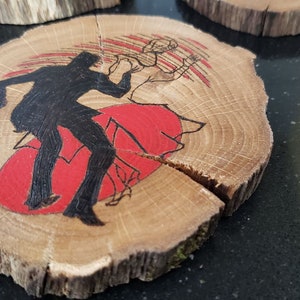 May include: Wooden coasters with a rustic, natural edge. Each coaster features a black silhouette of a dancing couple over a red backdrop. The wood grain is visible, adding to the handmade aesthetic.