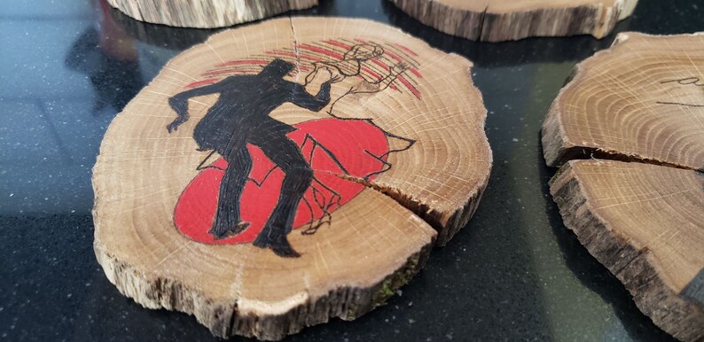 May include: Wooden coasters with a rustic, natural edge. Each coaster features a black silhouette of a dancing couple over a red backdrop. The wood grain is visible, adding to the handmade aesthetic.