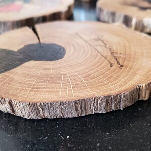 May include: Wooden coasters made from tree cross-sections. The coasters have a natural wood grain pattern with a dark, possibly stained, area. Some coasters have a signature. The edges are rough and unfinished.