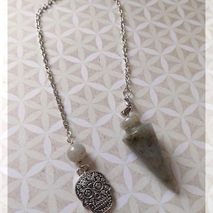 Może przedstawiać: Srebrny łańcuszek z szarym kryształowym wahadłem i srebrnym wisiorkiem z detalicznym wzorem czaszki cukrowej. Łańcuszek wisi na białym tle z geometrycznym wzorem.