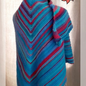 Peut inclure: Un châle crocheté à rayures bleues, violettes et rouges drapé sur un mannequin blanc. Le châle est réalisé avec un point chevron. Le mot "Flaure" est écrit en noir en haut de l'image.