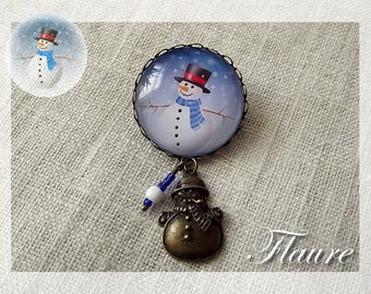 May include: A snowman charm with a blue scarf and black hat, surrounded by a white and blue glass dome. The charm is attached to a bronze snowman pendant with a blue and white beaded chain.