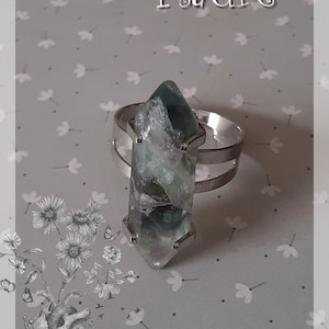 Peut inclure: Une bague en argent avec une pointe de cristal verte et blanche. La bague est sur un fond gris avec un motif floral.