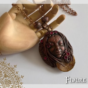 Puede incluir: Un collar marrón y cobre con un colgante que presenta una cara esculpida con los ojos cerrados y acentos rosados. El colgante está rodeado por un diseño texturizado, marrón y cobre.