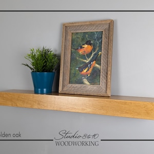May include: A golden oak floating shelf with a framed bird painting and a small potted plant. The painting features two orange and black birds perched on a branch. The shelf is mounted on a gray wall. The text "golden oak" and "Studio 8 to 10 WOODWORKING" are visible.