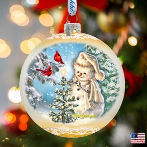 May include: A white glass Christmas ornament with a painted scene of a snowman standing next to a small Christmas tree with two red cardinals perched on a branch. The snowman is wearing a white hat and scarf and is holding a star on top of the tree. The ornament has a gold trim and the words "Dona Gelsinger" are written in the bottom right corner.