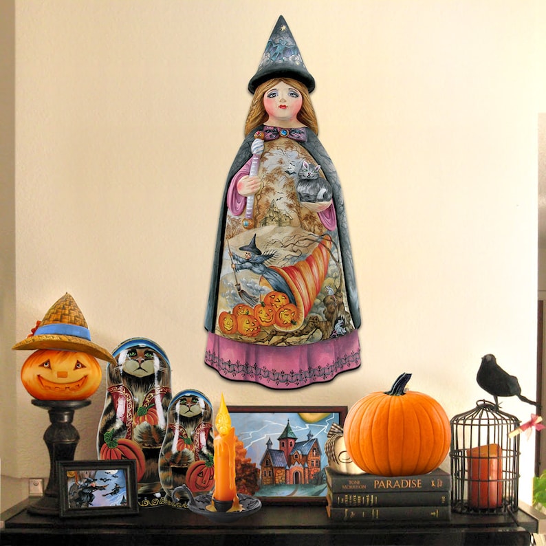 Peut inclure: Une figurine de sorci&egrave;re d'Halloween en bois avec une robe noire et rose, un chapeau pointu et un chat dans ses bras. La figurine est entour&eacute;e de citrouilles, d'un chat et d'un oiseau noir. La figurine est sur une &eacute;tag&egrave;re avec d'autres d&eacute;corations d'Halloween.