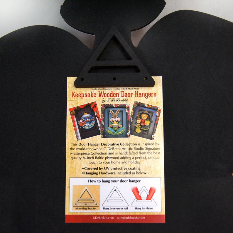 May include: A white card with a black border and text that reads "Keepsake Wooden Door Hangers by G.DeBrekht". The card shows three images of wooden door hangers with different holiday themes. The text also includes instructions on how to hang the door hangers.