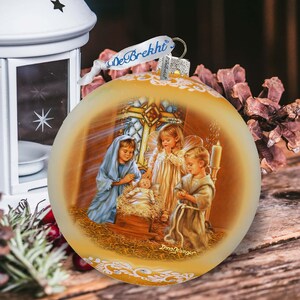 May include: A round Christmas ornament with a religious scene. The ornament has a gold-colored background with a painting of the Nativity. The scene includes Mary, angels, and baby Jesus. The ornament has a ribbon that says "DeBrekht".